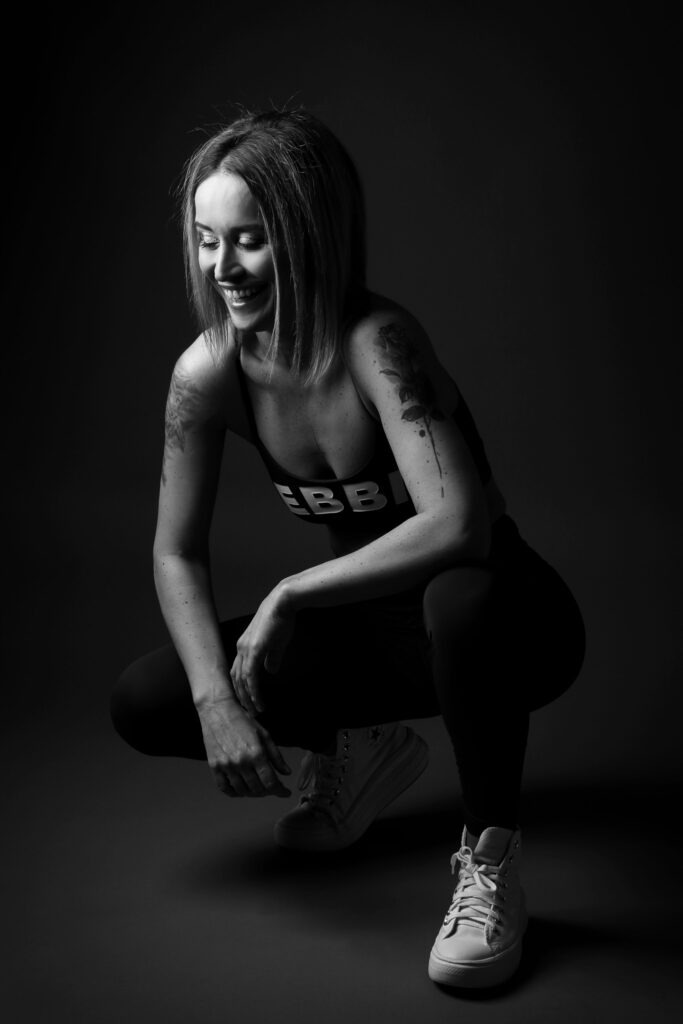Černobílá sportovní fotografie usměvavé ženy v podřepu na tmavém ateliérovém pozadí.